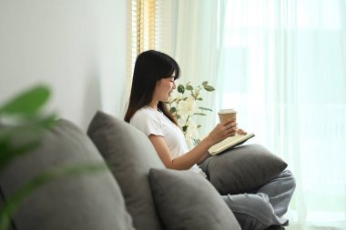 Evdeki kanepede bir fincan kahve içip kitap okuyan barışçıl genç bir kadın..