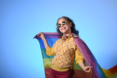 Güneş gözlüklü neşeli orta yaşlı kadın mavi arka planda gökkuşağı bayrağı taşıyor. LGBT, insan hakları ve eşitlik kavramı.