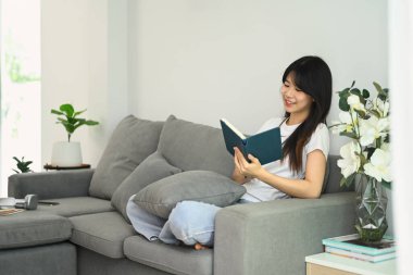 Evdeki kanepede günlük giysiler içinde kitap okuyan mutlu genç bir kadın..