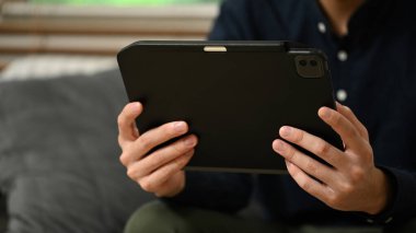 Genç işadamı dijital tablette e-posta okuyor ve oturma odasında kanepede dinleniyor..