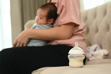 Bir bebek şişesi ahşap yemek masasında çalışan anne ve bebeğe karşı arka planda oturuyor..