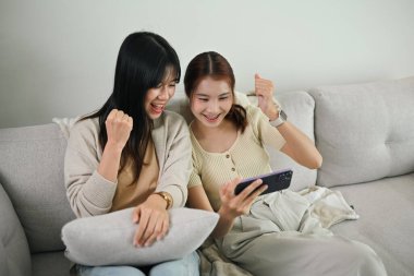 İki neşeli genç Asyalı kadın akıllı telefondan güzel sürpriz haberler okuyor ve evet hareketiyle kutluyorlar..