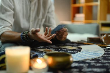 Birkaç boncuklu bilezik takan bir falcının yakın plan fotoğrafı. Çeşitli ruhsal öğelerin yanında tarot kartı tutuyor..