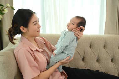 Genç Asyalı annenin bebeğini nazikçe kanepede otururken çekmesi. Çocuk bakımı ve annelik kavramı.
