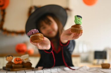 Cadı kostümlü tatlı kız elinde lezzetli Cadılar Bayramı kekleri tutuyor. Eldeki odaklanmayı seç.
