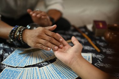 Kapalı çekim falcısı boncuklu bilezikler takıyor, masada çeşitli manevi eşyalarla el çizgileri okuyor..