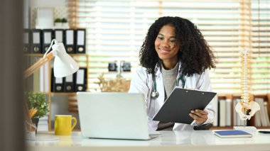 Beyaz önlüklü gülümseyen afro kadın doktor hastalarına uzaktan danışıyor..