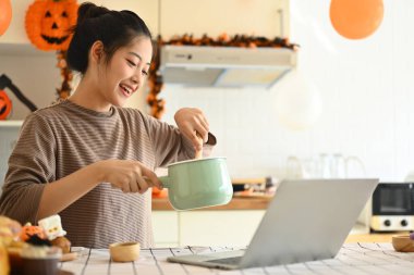 Mutlu genç bir kadın yemek pişiriyor ve Cadılar Bayramı için dekore edilmiş bir mutfakta dizüstü bilgisayarda tarifini takip ediyor..