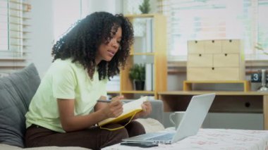 Genç Afro kadın serbest çalışan oturma odasında dizüstü bilgisayar kullanıyor ve deftere yazıyor..