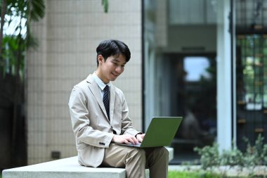 Takım elbiseli çekici bir işadamı toplantıdan önce bankta oturuyor ve laptopta e-postaları kontrol ediyor..