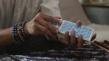 Yavaş çekimde, elinde mumların ve çeşitli ruhani eşyaların yanındaki tarot kartlarını karıştıran bir adam..
