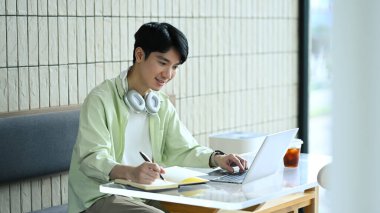 Genç adam modern bir kafede bir masada oturuyor dizüstü bilgisayarla çalışıyor ve notlar yazıyor..