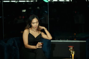 Gece elbisesi giymiş zarif genç bir kadın içki tutuyor loş bir salonda tek başına oturuyor. Gece hayatı ve lüks yaşam tarzı konsepti.