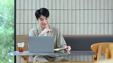 Modern bir kafede oturmuş laptopla çalışıp notlar yazan Asyalı hoş bir adam..