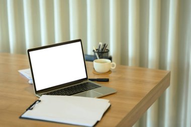 Beyaz boş ekranlı dizüstü bilgisayar, dosya, kahve fincanı ve ahşap ofis masasında kırtasiye malzemesi..