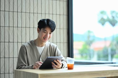 Gülümseyen Asyalı genç adam kahve dükkanında pencerenin yanında oturmuş kitap okuyor..