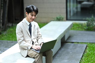 Açık bej takım elbiseli gülümseyen iş adamı beton bir bankta oturuyor ve dizüstü bilgisayar kullanıyor..