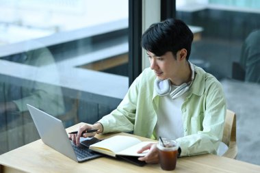 Yakışıklı Asyalı adam dizüstü bilgisayar kullanıyor ve not alıyor. İnsanlar, teknoloji ve uzaktan çalışma konsepti.