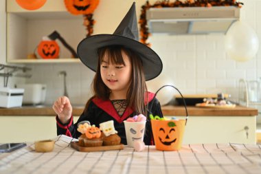 Cadılar Bayramı 'nda cadı gibi giyinmiş küçük kız mutfak masasında farklı canavarlarla kek süslüyor..