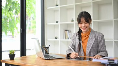 Modern bir ofiste masasında dijital tablet kullanan gülümseyen genç bir iş kadını..
