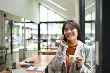 Neşeli genç yaratıcı kadın kahve içiyor ve modern ofiste cep telefonuyla konuşuyor..