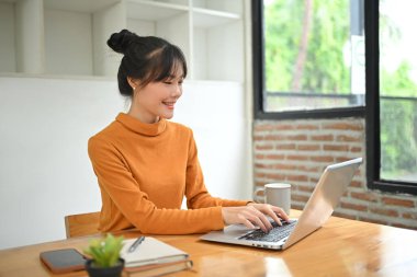 Ofiste dizüstü bilgisayar kullanarak turuncu balıkçı yaka kazak giyen gülümseyen genç Asyalı kadın..