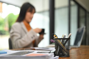 Ofis malzemelerinin ve mali raporların resmi ahşap masada, iş kadını arka planda oturuyor..