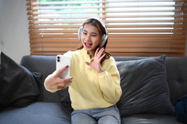 Neşeli genç Asyalı kadın oturma odasındaki kanepede oturmuş akıllı telefondan bir video görüşmesi yapıyor..