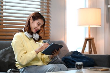 Evde dijital tablet tasarım projesinde çalışan, kulaklıklı, gülümseyen genç bir kadın..