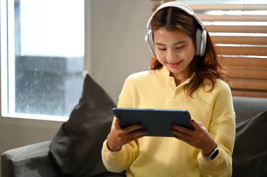 Kulaklık takan güzel bir Asyalı kadın evde dijital tabletten video izliyor..