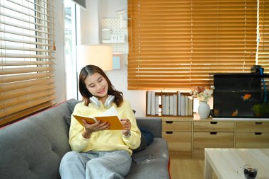 Gülümseyen genç bir kadın oturma odasında dinleniyor ve kitap okuyor, evde boş zamanların keyfini çıkarıyor..