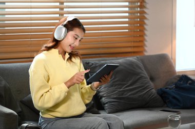 Kablosuz kulaklık takan, evdeki dijital tablette internette sörf yapan çekici genç bir kadın..