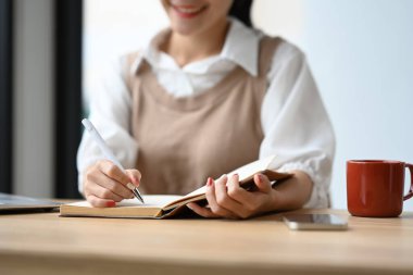 Gülümseyen genç bir kadın defter yazıyor, ofis masasında kağıt notlar üzerinde fikirler planlıyor..