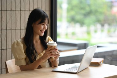 Elinde bir fincan kahve tutan ve bilgisayarında iş e-postaları okuyan çekici genç bir kadın..