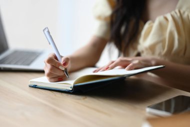 Gülümseyen genç bir kadın defter yazıyor, ofis masasında kağıt notlar üzerinde fikirler planlıyor..