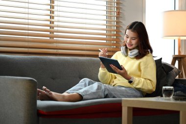 Oturma odasındaki kanepede dijital tablet kullanan genç ve güzel bir kadın..