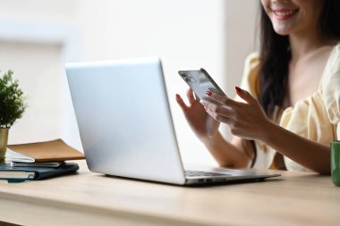 Çalışma masasında dizüstü bilgisayarıyla gülümseyen ve cep telefonu kullanan genç bir kadın..