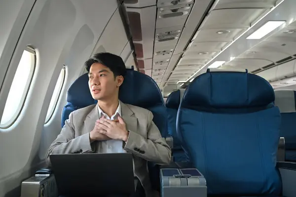 stock image Young businessman looking out the window with hand resting on his chest, experiencing aerophobia or fear of flying.