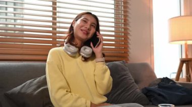 Gülümseyen genç Asyalı kadın evdeki kanepede otururken telefonla konuşuyor..