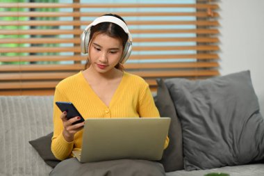 Kulaklık takan genç ve güzel Asyalı bir kadın dizüstü bilgisayarla koltukta oturuyor ve cep telefonu kullanıyor..