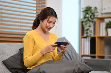 Genç, güzel Asyalı kadın evdeki kanepede dinleniyor ve akıllı telefonuyla video oyunları oynuyor..