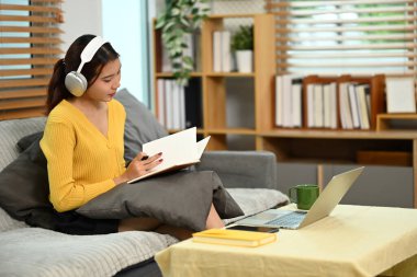 Kulaklıklı genç kadın evdeki kanepede dizüstü bilgisayar kullanıyor. Uzak çalışma ve çevrimiçi öğrenme kavramı.