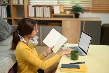 Elinde not defteri olan ve oturma odasında dizüstü bilgisayarla çalışan odaklanmış bir kız öğrenci..