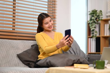 Mutlu genç bir kadın evdeki kanepede akıllı telefon kullanarak arkadaşlarıyla sohbet etmekten hoşlanıyor..