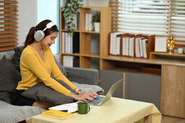 Genç bir kadın kanepeye oturmuş kulaklık takıyor ve laptopuyla çalışıyor..