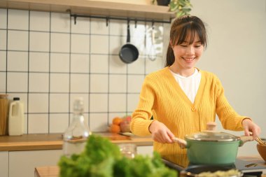 Gülümseyen genç kadın mutfak içinde yemek pişirme sürecinin keyfini çıkarıyor..