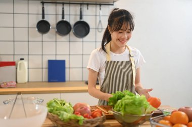 Önlük giymiş gülümseyen genç Asyalı kadın mutfakta taze ve sağlıklı bir salata hazırlıyor..