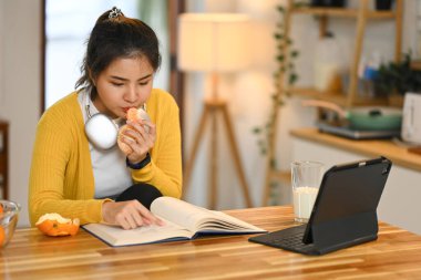 Güzel genç Asyalı kadın mutfak tezgahında portakal yerken kitap okuyor..
