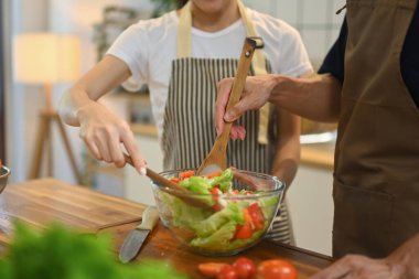 Önlük giymiş genç bir çiftin ahşap tezgahta taze bir salata hazırlarken çekilmiş görüntüsü..