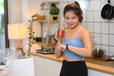 Çekici, sportif, genç bir kadın. Antrenmandan sonra sıcak çayla dinleniyor ve saatini kontrol ediyor..